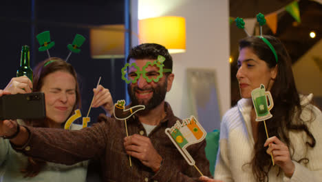 Group-Of-Friends-Dressing-Up-At-Home-Or-In-Bar-Celebrating-At-St-Patrick's-Day-Party-Posing-For-Selfie-On-Phone-2